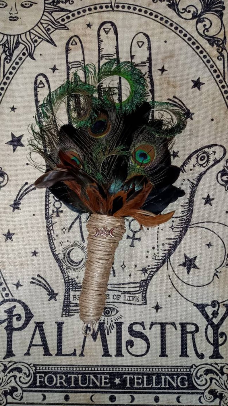 May include: A peacock feather fan with a brown leather handle and a triple moon charm. The fan is set against a background of a palmistry chart with stars and moons.