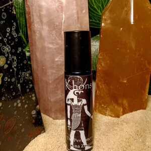 May include: A black roll-on bottle with a white label featuring an Egyptian goddess design and the text "Khonsu The Perfumers of Luna Zugar", surrounded by pink and orange crystals.