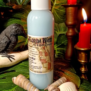May include: A blue glass bottle with a white spray nozzle and a label that reads "Florida Water by Luna Zingara". The label features a map of Florida and text about the product's ingredients and uses. The bottle is sitting on a green leafy surface with seashells.