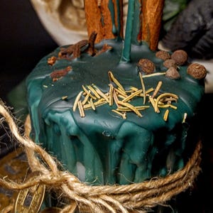 May include: A dark teal candle with cinnamon sticks, herbs, and spices. The candle is wrapped with twine and adorned with gold coins. The candle has a rustic, natural aesthetic.