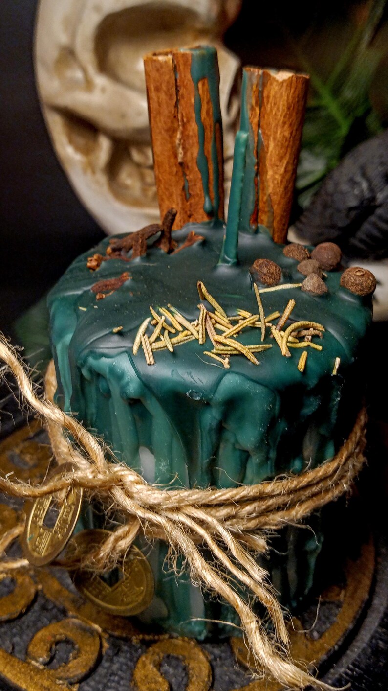 May include: A dark teal candle with cinnamon sticks, herbs, and spices. The candle is wrapped with twine and adorned with gold coins. The candle has a rustic, natural aesthetic.