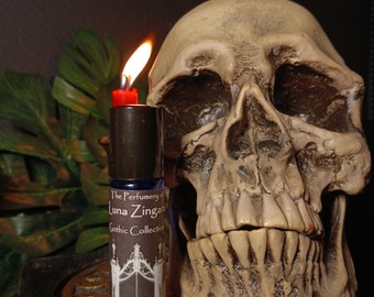 May include: A blue glass bottle with a black label that reads "The Perfumery of Luna Zingara Gothic Collection Blood Moon". The bottle is sitting in front of a white skull with a toothy grin.