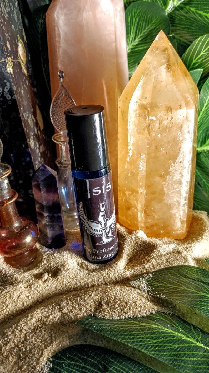 May include: A dark blue rollerball perfume bottle with the word "Isis" and an illustration of a winged figure. The bottle is surrounded by crystals, including a large orange quartz and a pink crystal, on a bed of sand.