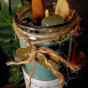May include: A teal green glass jar candle with a white label that reads "Dust Woman Energy & Abundance Ritual Candle". The jar is tied with twine and decorated with a gold coin charm. The candle is surrounded by various ingredients, including cinnamon sticks, a dried leaf, and a green crystal.