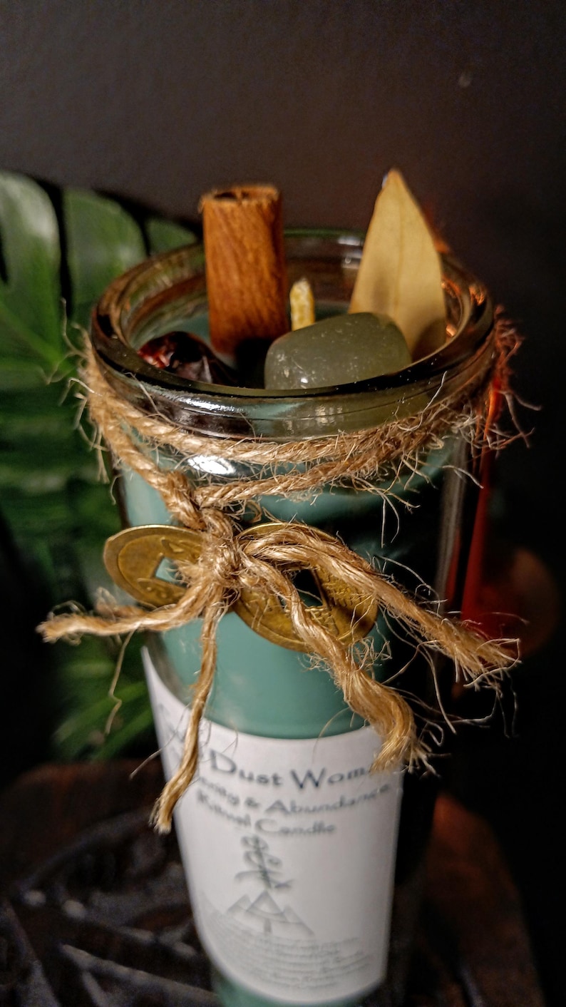 May include: A teal green glass jar candle with a white label that reads "Dust Woman Energy & Abundance Ritual Candle". The jar is tied with twine and decorated with a gold coin charm. The candle is surrounded by various ingredients, including cinnamon sticks, a dried leaf, and a green crystal.