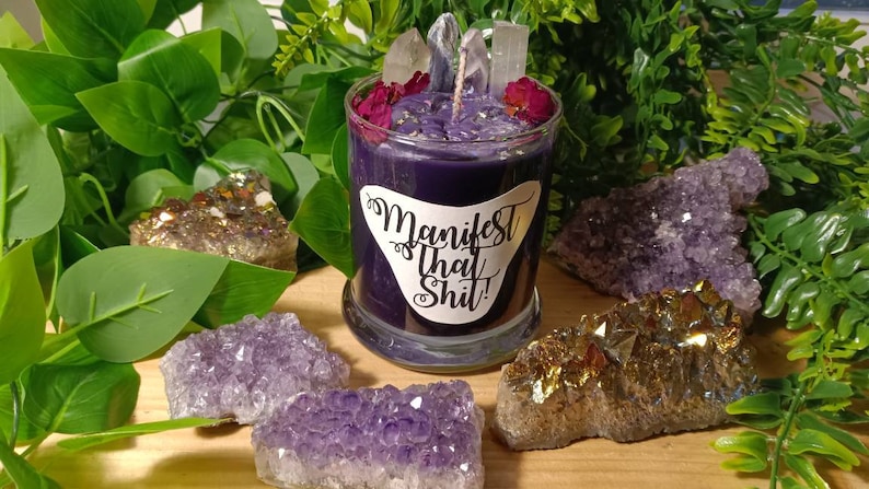 May include: Purple candle in a glass jar with a white label that says "Manifest That Shit!" The candle is surrounded by amethyst and citrine crystals.