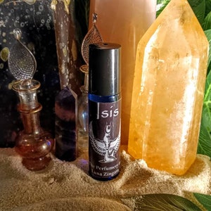 May include: A blue glass roller bottle labeled "Isis" with a black cap, featuring a winged figure design. The bottle is surrounded by crystals, a decorative glass bottle, and greenery, creating an aesthetic display.
