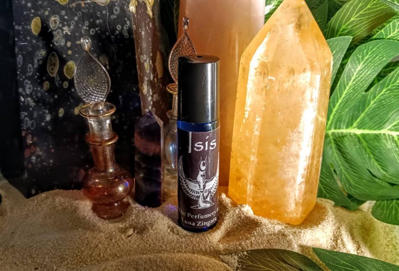 May include: A blue glass roller bottle labeled "Isis" with a black cap, featuring a winged figure design. The bottle is surrounded by crystals, a decorative glass bottle, and greenery, creating an aesthetic display.