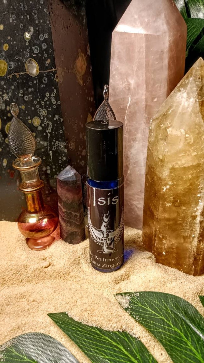 May include: A dark brown roll-on bottle with the word "Isis" in white, featuring a winged figure design. The bottle is surrounded by crystals and a small glass bottle, set on a sandy surface with green leaves.