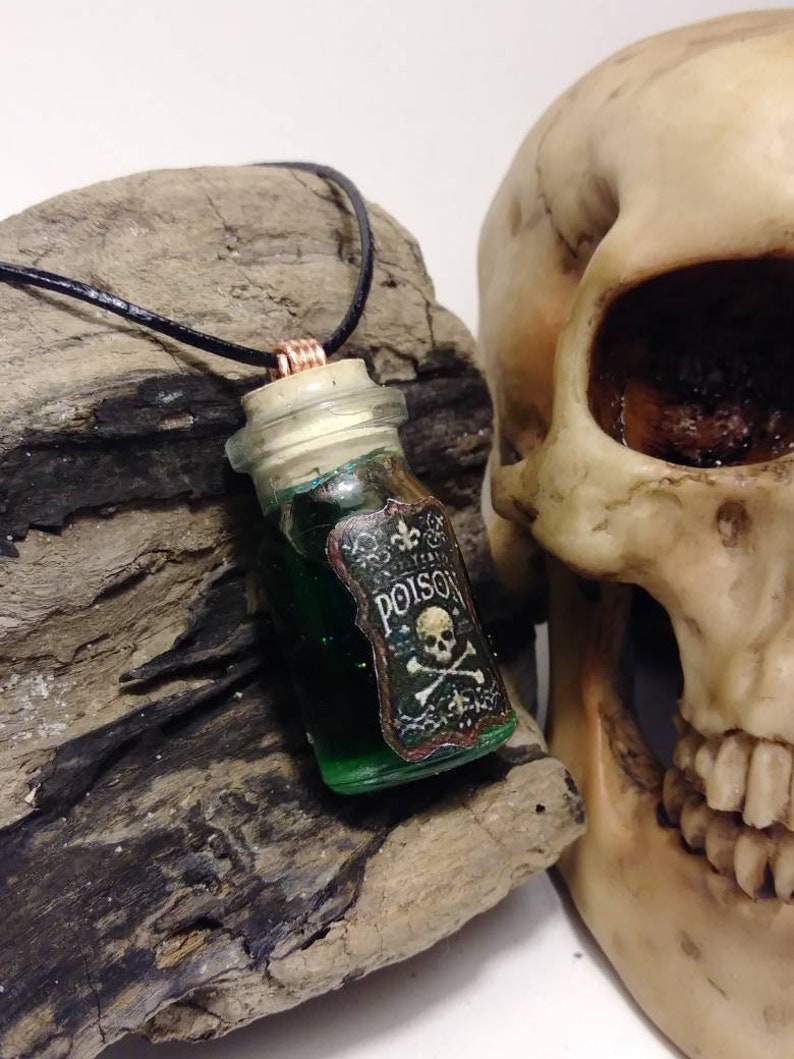 May include: A green glass bottle pendant necklace with a cork stopper and a label that reads "Poison" with a skull and crossbones. The necklace is on a black cord and is displayed next to a white skull.