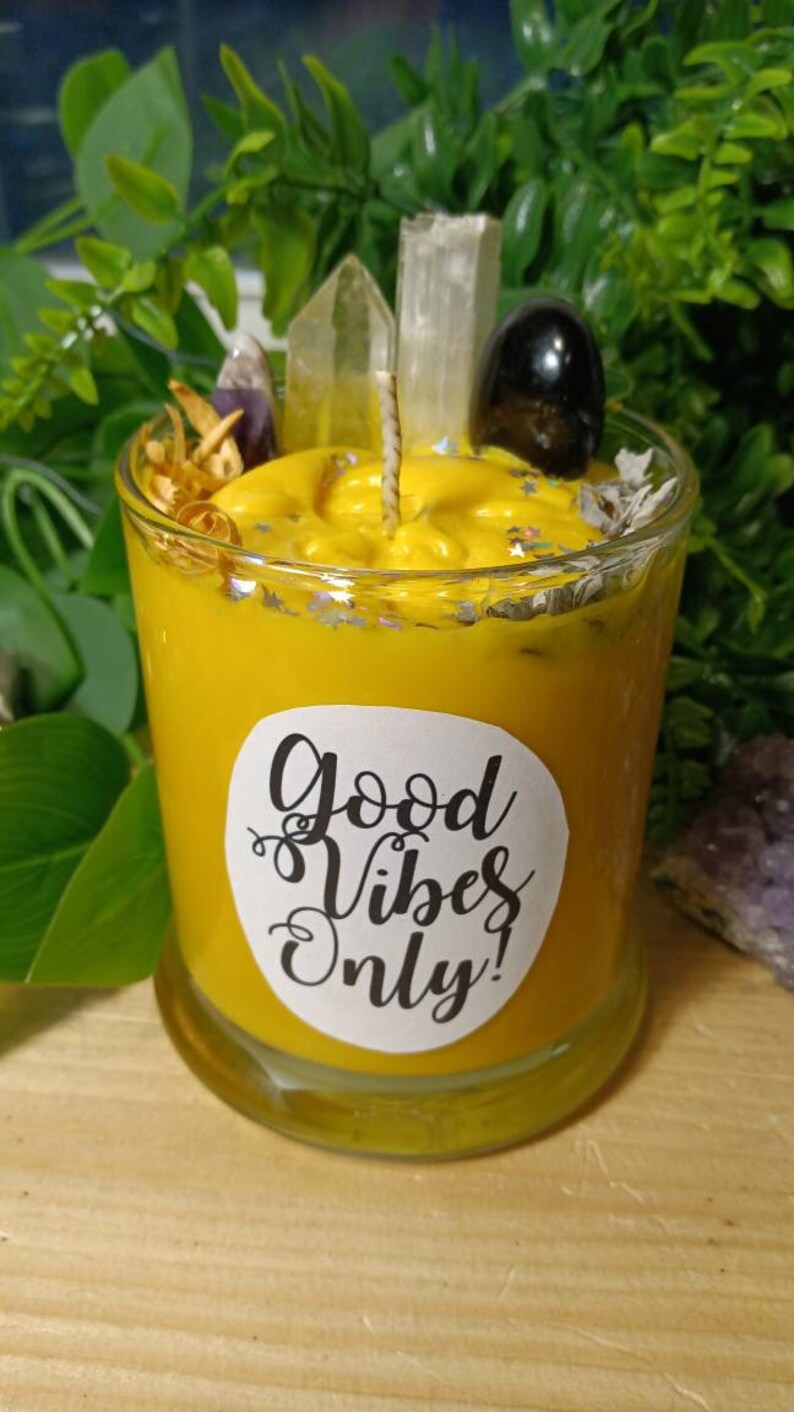 May include: A yellow candle in a glass jar with a white label that says "Good Vibes Only!" The candle is surrounded by crystals and dried herbs.