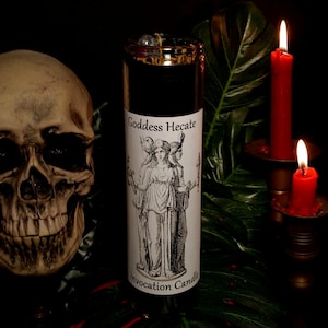 May include: A black candle with a white label that reads "Goddess Hecate Invocation Candle". The label features a black and white illustration of a woman with three heads. The candle is surrounded by two red candles in candle holders.