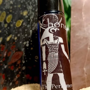 May include: A brown glass roller bottle with a black cap. The bottle has a white image of an Egyptian god with a falcon head and the text "The Perfume of Luna Zima" on the label.