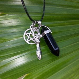 May include: A silver pendant necklace with a black crystal point, a pentagram, and a silver wand charm. The necklace is on a black cord and is resting on a green leaf.