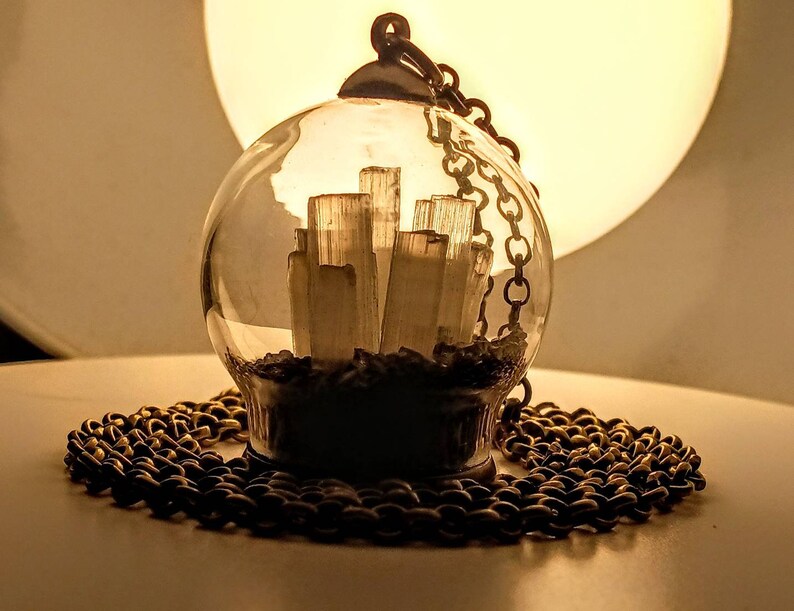 Peut inclure: Un collier avec un pendentif en forme de globe en verre et une cha&icirc;ne dor&eacute;e. &Agrave; l'int&eacute;rieur du globe se trouvent des cristaux de s&eacute;l&eacute;nite blancs.