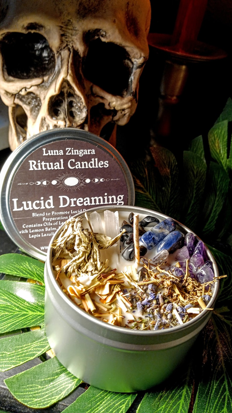May include: A silver tin candle with a black label that reads "Luna Zingara Ritual Candles Lucid Dreaming". The candle is filled with white wax, dried herbs, and crystals.