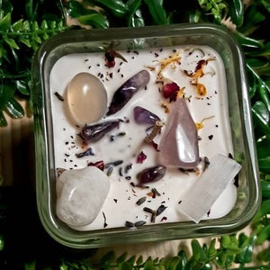 Pode incluir: Uma vela em um pote de vidro quadrado com uma base branca e v&aacute;rios cristais incrustados na cera. Os cristais incluem quartzo claro, ametista, quartzo rosa e selenita. A vela &eacute; cercada por folhagem verde.