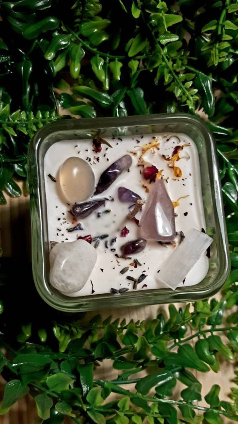 Pode incluir: Uma vela em um pote de vidro quadrado com uma base branca e v&aacute;rios cristais incrustados na cera. Os cristais incluem quartzo claro, ametista, quartzo rosa e selenita. A vela &eacute; cercada por folhagem verde.