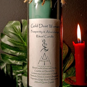 May include: A teal green glass candle with a white label that reads "Gold Dust Woman Prosperity & Abundance Ritual Candle". The label also includes a list of herbs, crystals, and sacred oils used in the candle. The candle is tied with twine and has two gold coins attached.