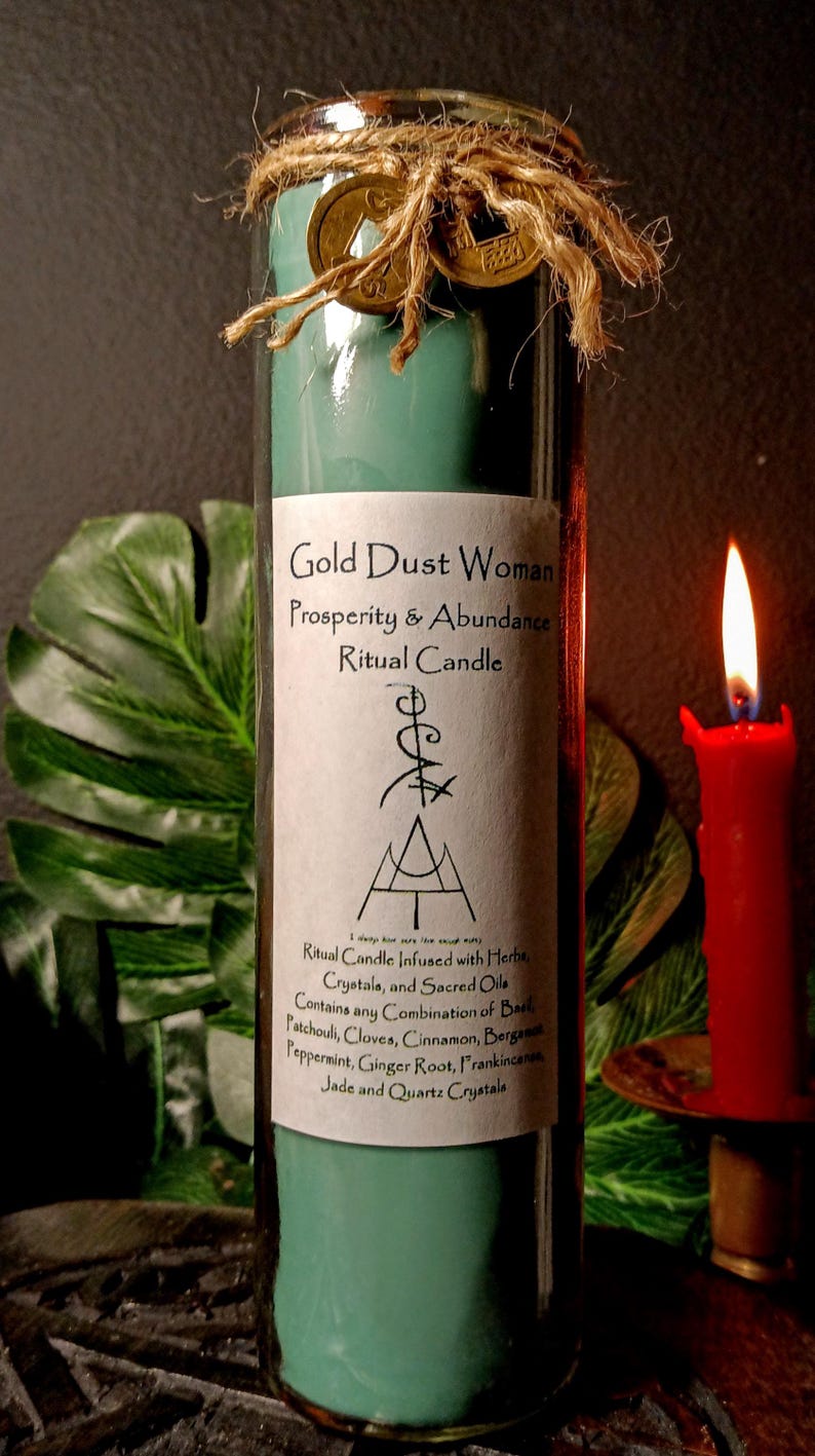 May include: A teal green glass candle with a white label that reads "Gold Dust Woman Prosperity & Abundance Ritual Candle". The label also includes a list of herbs, crystals, and sacred oils used in the candle. The candle is tied with twine and has two gold coins attached.