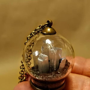 Peut inclure: Un pendentif en forme de globe en verre transparent contenant un amas de cristal de s&eacute;l&eacute;nite argent&eacute;, avec une cha&icirc;ne et un fermoir en laiton.