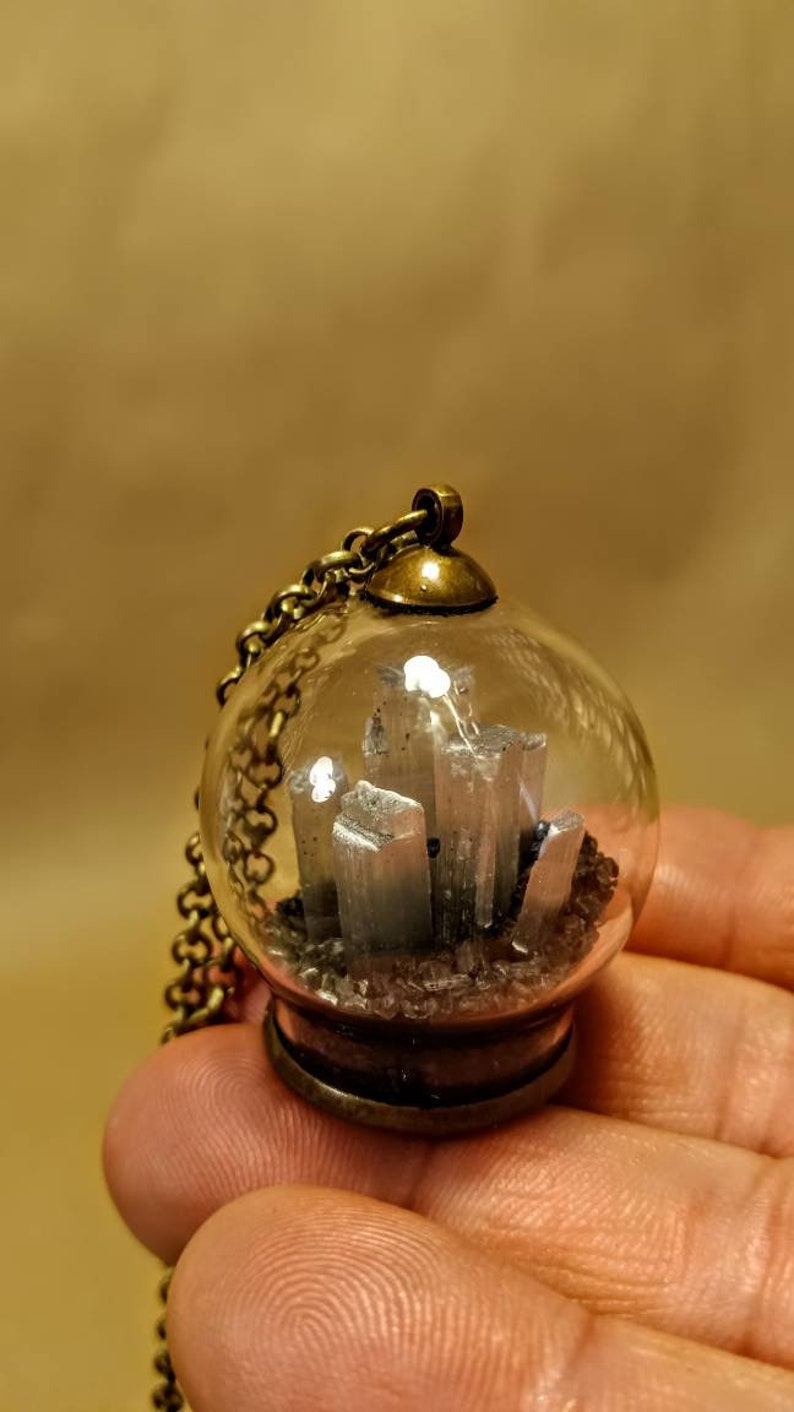 Peut inclure: Un pendentif en forme de globe en verre transparent contenant un amas de cristal de s&eacute;l&eacute;nite argent&eacute;, avec une cha&icirc;ne et un fermoir en laiton.