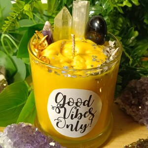 May include: A yellow candle with a white label that says "Good Vibes Only!" The candle is surrounded by crystals and other decorative elements.