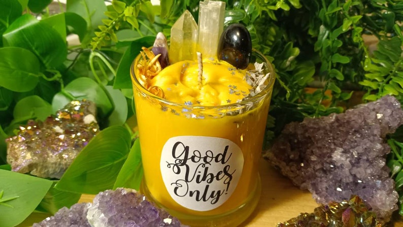 May include: A yellow candle with a white label that says "Good Vibes Only!" The candle is surrounded by crystals and other decorative elements.