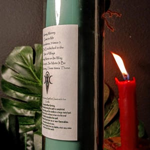 May include: A teal green glass candle with a white label that reads "Money Money Come to Me Abundance, 3 times 3. Be Enriched in the Best of Ways.  Let None on Its Way Accept, So Mote It Be. Money Three times Three."  The candle is wrapped in twine and has a wooden top.