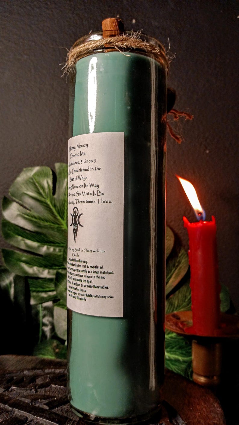 May include: A teal green glass candle with a white label that reads "Money Money Come to Me Abundance, 3 times 3. Be Enriched in the Best of Ways.  Let None on Its Way Accept, So Mote It Be. Money Three times Three."  The candle is wrapped in twine and has a wooden top.