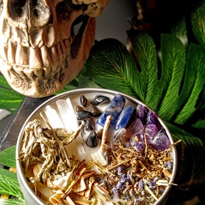 May include: A white candle with various herbs, crystals, and stones embedded in the wax. The candle is in a silver tin and is surrounded by green leaves.