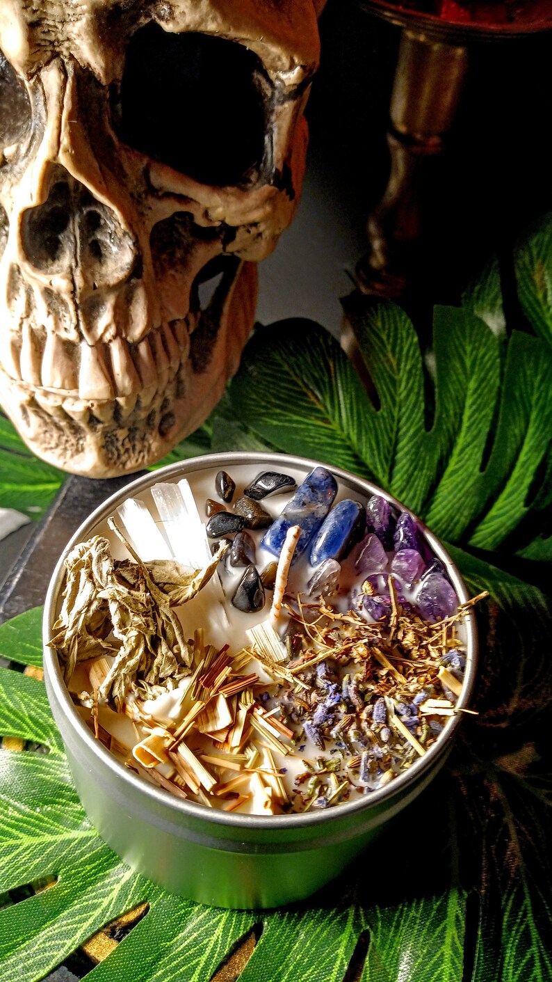 May include: A white candle with various herbs, crystals, and stones embedded in the wax. The candle is in a silver tin and is surrounded by green leaves.