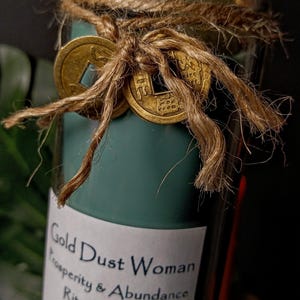 May include: A teal green glass candle with a white label that reads "Gold Dust Woman Prosperity & Abundance Ritual Candle". The candle is decorated with gold coins and twine.