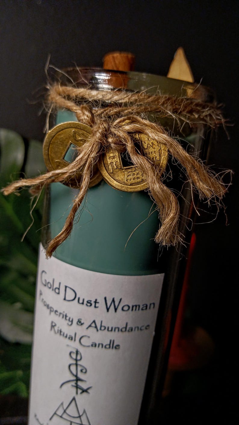 May include: A teal green glass candle with a white label that reads "Gold Dust Woman Prosperity & Abundance Ritual Candle". The candle is decorated with gold coins and twine.