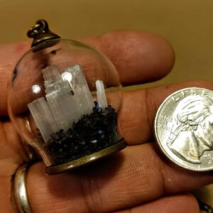 Peut inclure: Un collier avec un pendentif en verre contenant un petit globe en verre transparent rempli de cristaux de s&eacute;l&eacute;nite blancs et de sable noir. Le pendentif est attach&eacute; &agrave; une cha&icirc;ne en bronze.