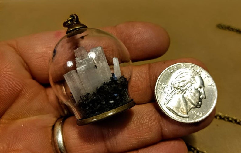 Peut inclure: Un collier avec un pendentif en verre contenant un petit globe en verre transparent rempli de cristaux de s&eacute;l&eacute;nite blancs et de sable noir. Le pendentif est attach&eacute; &agrave; une cha&icirc;ne en bronze.