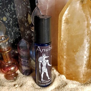 Puede incluir: Un frasco de vidrio azul con tapa negra y etiqueta. La etiqueta presenta una ilustraci&oacute;n en blanco y negro de Anubis, el dios egipcio de la muerte, y el texto "Anubis The Perfumer Luna Zingaro".