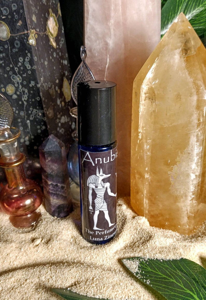 Puede incluir: Un frasco de vidrio azul con tapa negra y etiqueta. La etiqueta presenta una ilustraci&oacute;n en blanco y negro de Anubis, el dios egipcio de la muerte, y el texto "Anubis The Perfumer Luna Zingaro".