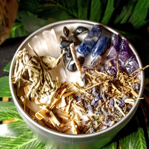 May include: A white candle with various crystals, herbs, and dried flowers embedded in the wax. The crystals include amethyst, clear quartz, and blue sodalite. The herbs include dried lavender and sage. The candle is in a silver tin.
