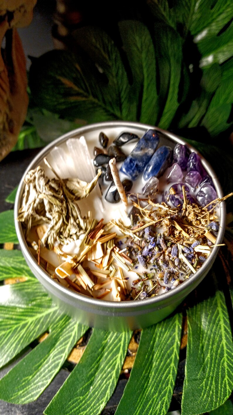 May include: A white candle with various crystals, herbs, and dried flowers embedded in the wax. The crystals include amethyst, clear quartz, and blue sodalite. The herbs include dried lavender and sage. The candle is in a silver tin.