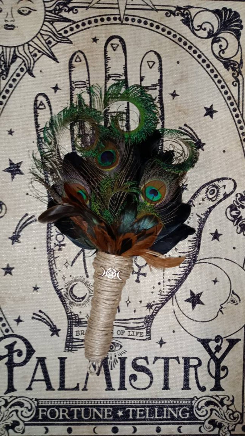 May include: A hand-tied bouquet of peacock feathers with a brown twine handle and a silver charm featuring a triple moon symbol. The bouquet is positioned in front of a vintage palmistry chart with the words "Palmistry Fortune Telling".