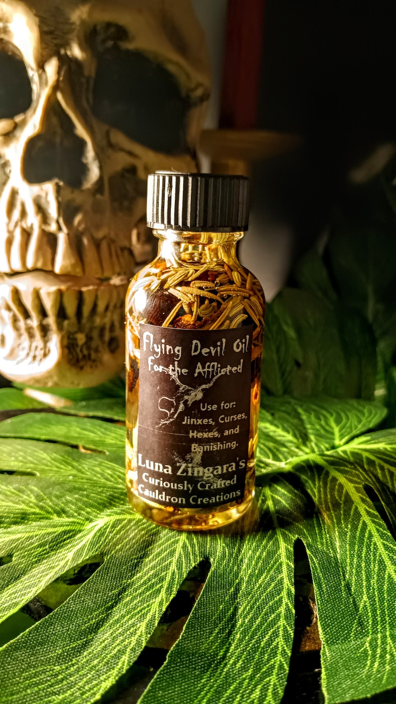 May include: A small glass bottle filled with a golden liquid and herbs. The label reads "Flying Devil Oil For the Afflicted" and "Luna Zingara's Curiously Crafted Cauldron Creations". The bottle is sitting on a green leaf.