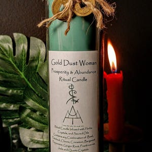 May include: A teal green glass candle with a gold label that reads "Gold Dust Woman Prosperity & Abundance Ritual Candle". The candle is decorated with gold coins and twine. The label also includes a symbol and a list of herbs, crystals, and sacred oils used in the candle.