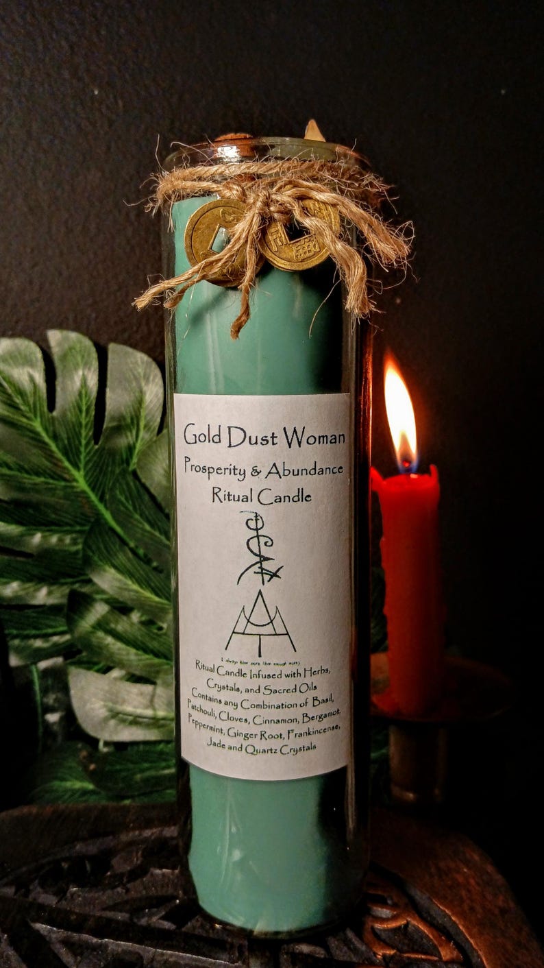 May include: A teal green glass candle with a gold label that reads "Gold Dust Woman Prosperity & Abundance Ritual Candle". The candle is decorated with gold coins and twine. The label also includes a symbol and a list of herbs, crystals, and sacred oils used in the candle.