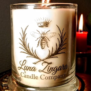 May include: A white candle in a clear glass jar with a gold bee and crown logo. The logo reads "Luna Zingara Candle Company".