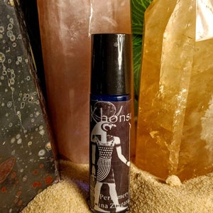 May include: A black roll-on bottle with a blue label featuring an illustration of an Egyptian god. The label also includes the text "The Perfumary of Luna Zingara". The bottle is sitting on a pile of sand.