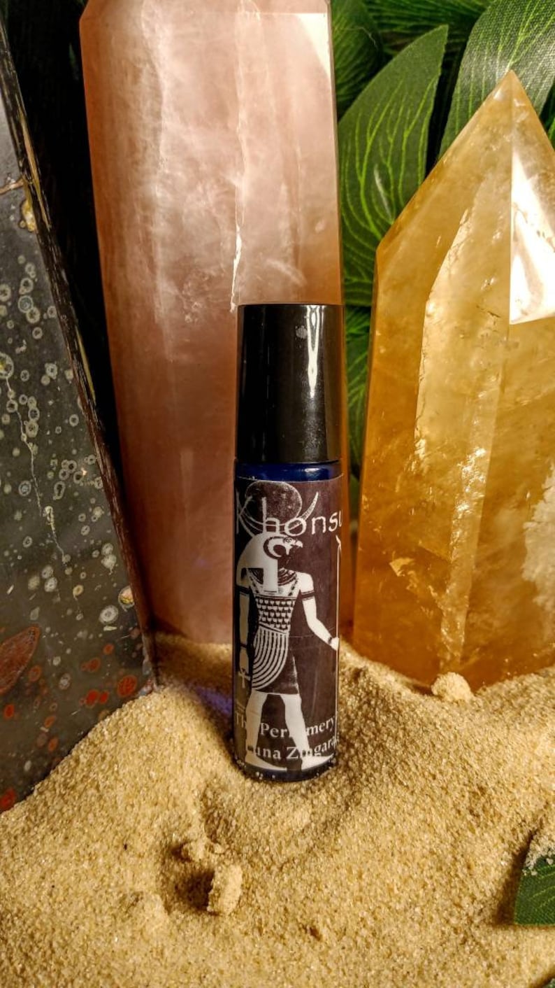 May include: A black roll-on bottle with a blue label featuring an illustration of an Egyptian god. The label also includes the text "The Perfumary of Luna Zingara". The bottle is sitting on a pile of sand.