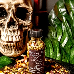 May include: A small glass bottle filled with herbs and labeled "Flying Devil Oil for the Afflicted". The label also includes instructions for use and the name "Luna Zingara's Curiously Crafted Cauldron Creations". The bottle is sitting on a dark surface with dried herbs and flowers.