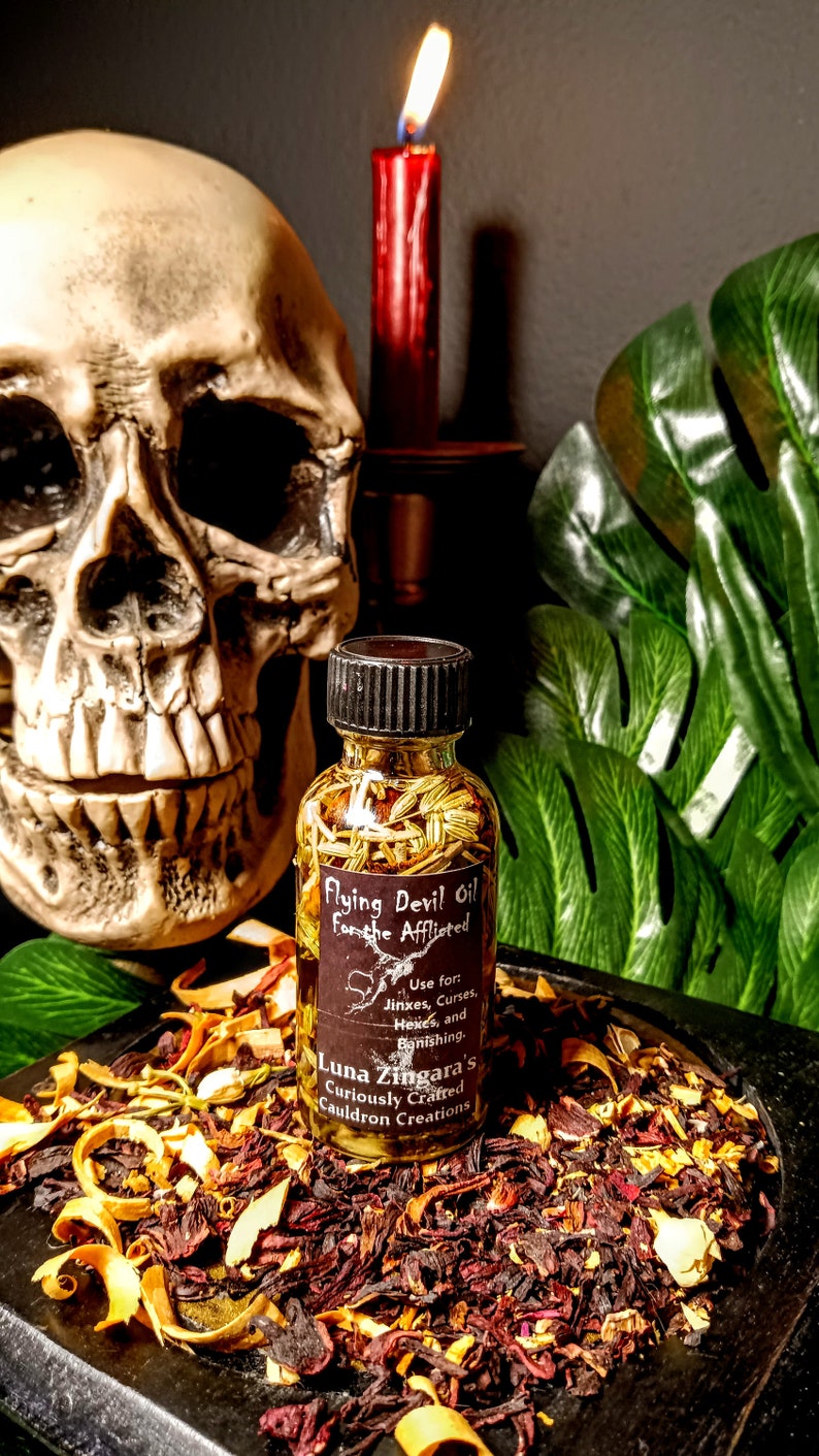May include: A small glass bottle filled with herbs and labeled "Flying Devil Oil for the Afflicted". The label also includes instructions for use and the name "Luna Zingara's Curiously Crafted Cauldron Creations". The bottle is sitting on a dark surface with dried herbs and flowers.