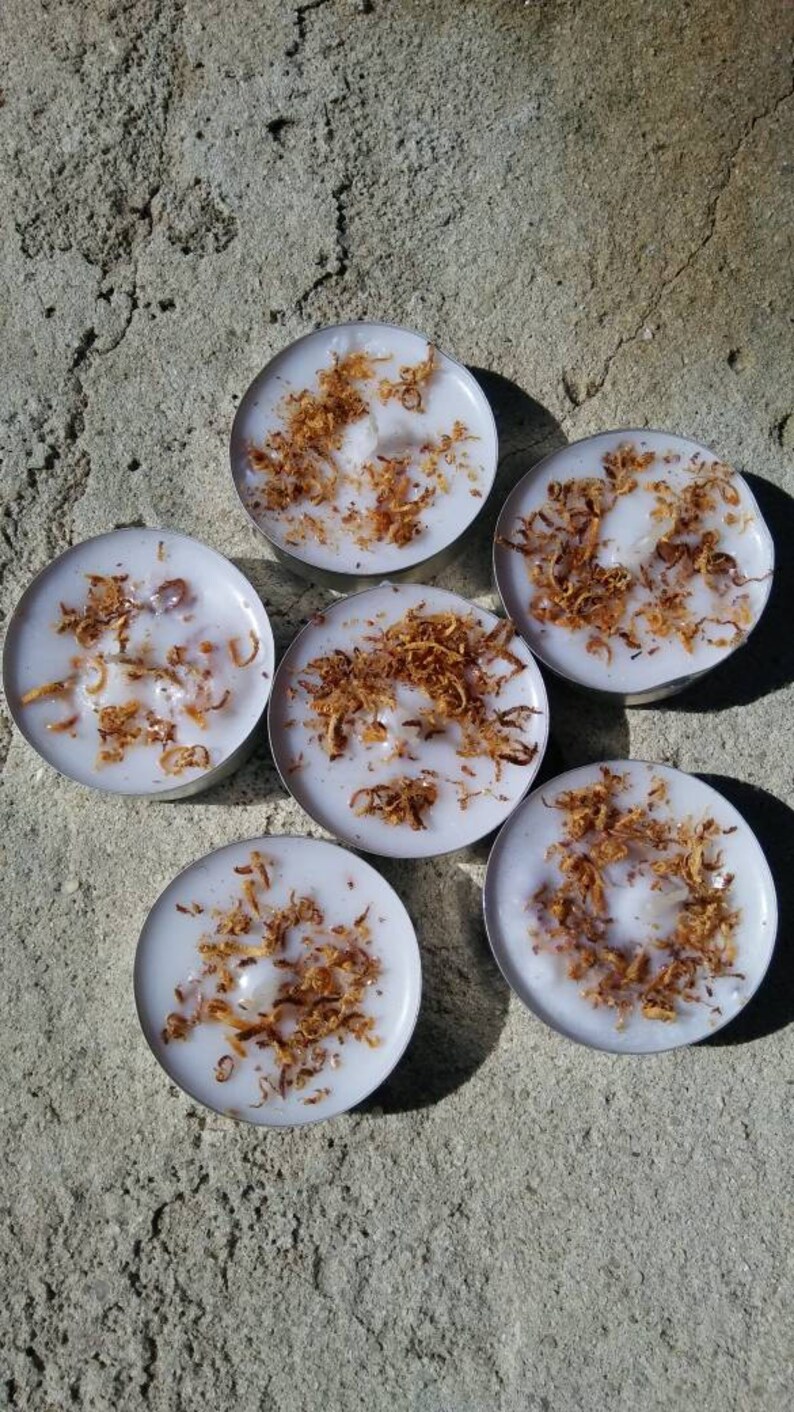 May include: Six white tea light candles with a brown, flaky topping. The candles are arranged in a circle on a gray concrete surface.